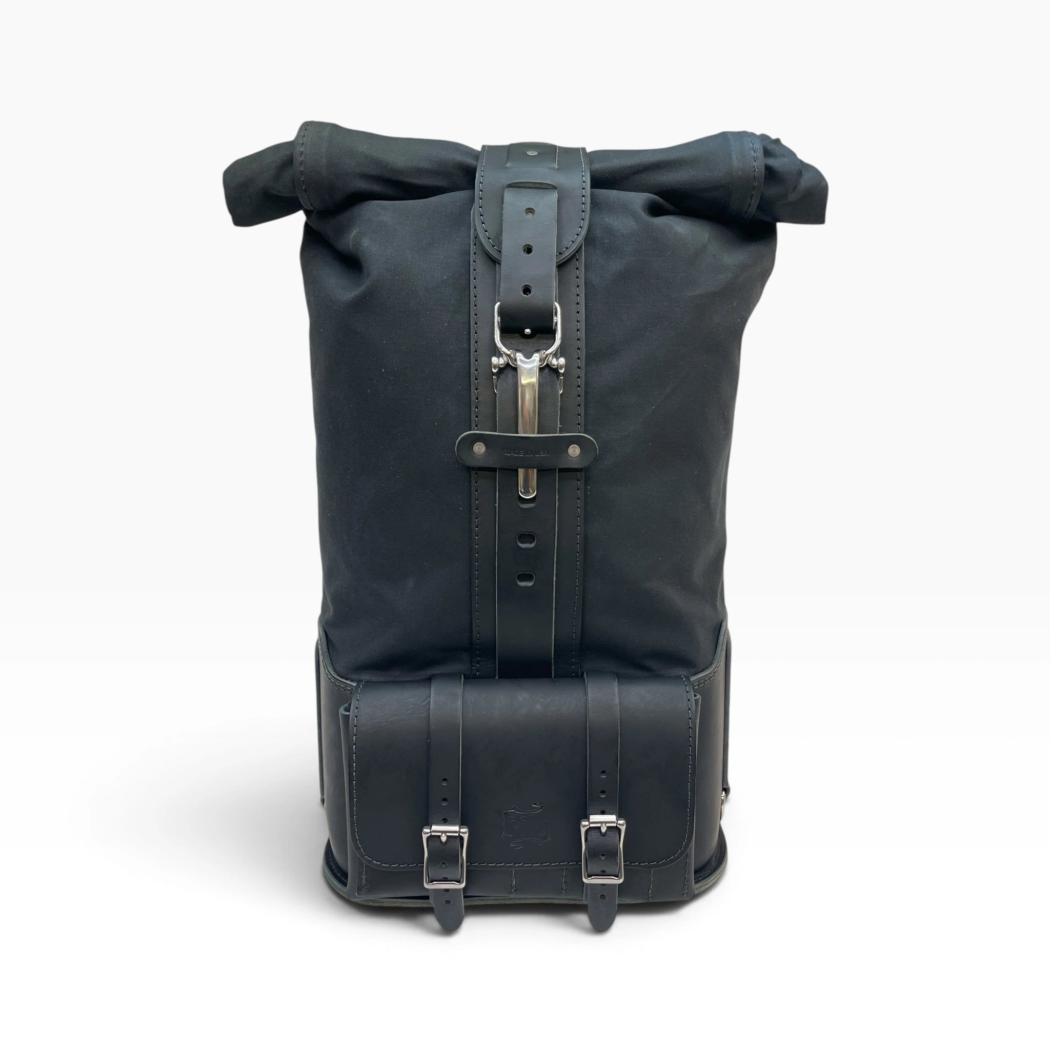 A black roll-top backpack with black leather straps, buckles, and a silver metal zipper pull, shown from the back against a light gray background.
