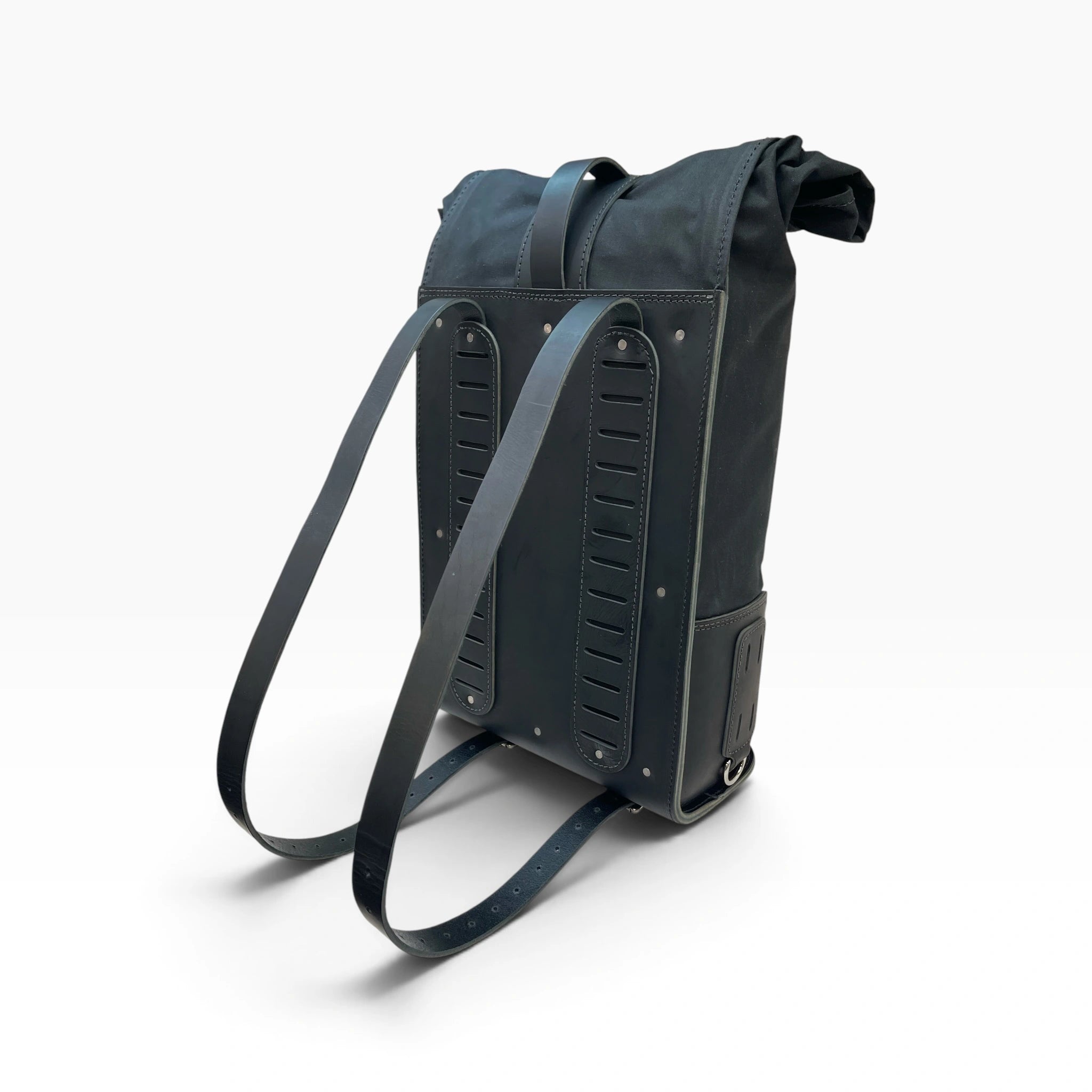 A black backpack shown from a three-quarters rear view, featuring a matching fabric shoulder strap and structured, ribbed leather-like side panels with metal rivets, set against a clean white background.